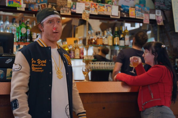 Un homme avec un papier dans le nez, visiblement sonné, se tient au bar avec une femme en rouge buvant un verre.