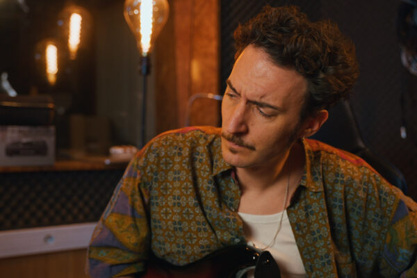 Un homme moustachu joue de la guitare dans un studio à l’éclairage tamisé.